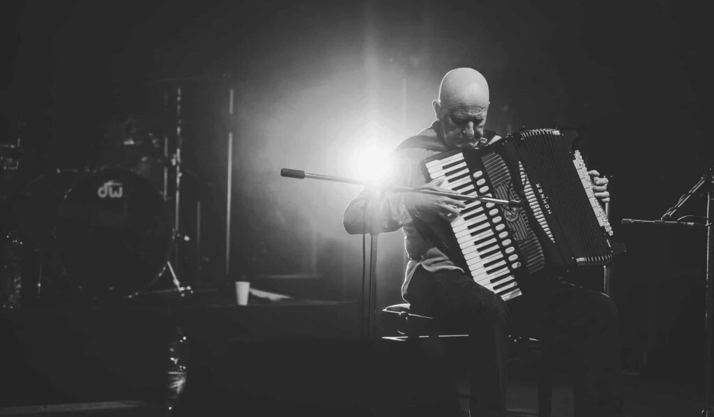Schwarz-Weiß-Foto eines glatzköpfigen Mannes auf der Bühne, der ein Akkordeon mit dramatischer Beleuchtung hinter ihm spielt. Im Hintergrund ist ein Schlagzeug zu sehen, das teilweise von Schatten und Rauch verdeckt wird - die perfekte Inspiration für jedes kreative Webdesign Aachen-Projekt.