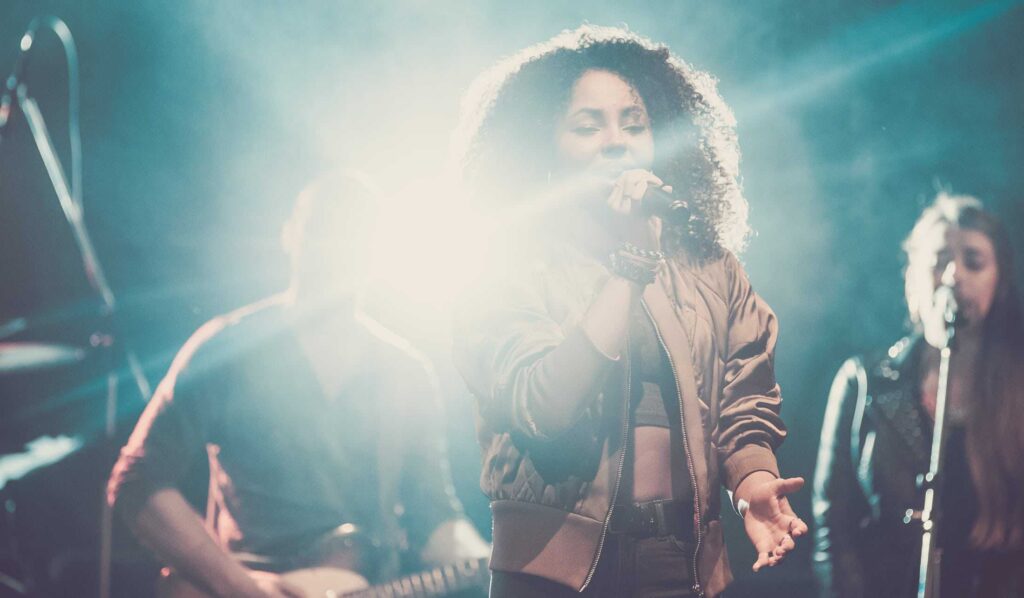 Eine Frau mit lockigem Haar singt auf der Bühne in ein Mikrofon, das von hellen Lichtern angestrahlt wird, während ihre Band im Hintergrund spielt - so wie die kreative Energie bei einer Veranstaltung der Webdesigner Aachen.