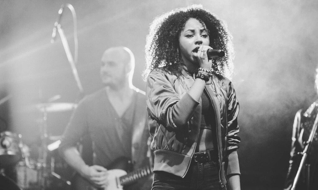 Eine Frau mit lockigen Haaren singt auf der Bühne in ein Mikrofon, sie trägt eine Jacke und Armbänder, im Hintergrund sind ein Gitarrist und ein Schlagzeug zu sehen - wie die kreative Energie bei einem Webdesign Aachen oder Wordpress Aachen Event.