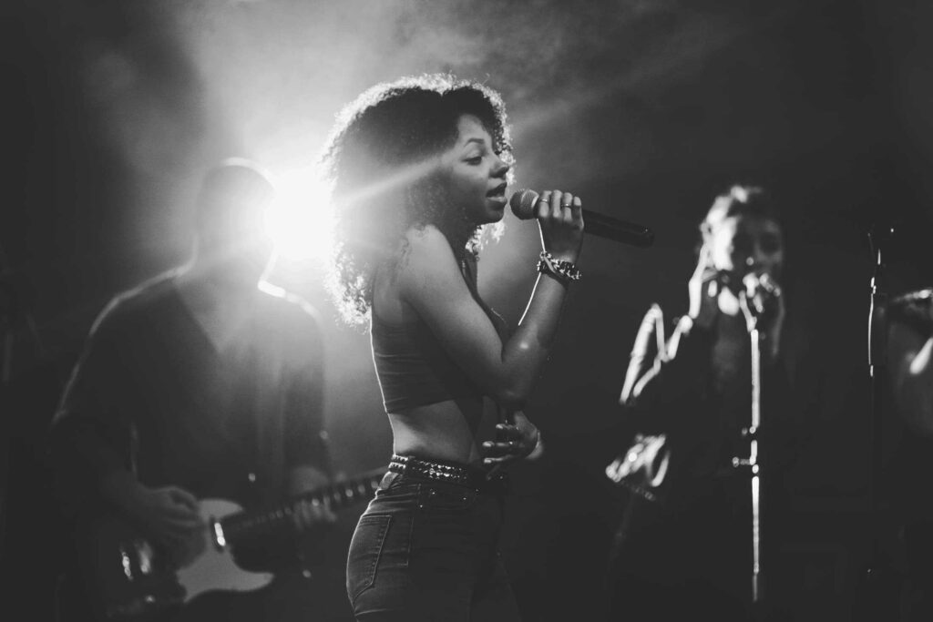 Eine Frau singt leidenschaftlich in ein Mikrofon auf der Bühne, umrahmt von hellen Lichtern. Zwei Musiker - einer an der Gitarre, ein anderer als Backgroundsänger - unterstützen sie im Hintergrund. Dieses Schwarz-Weiß-Bild fängt die Energie eines Live-Auftritts in Aachen ein, der Heimat des Top-Webdesigners Aachen.