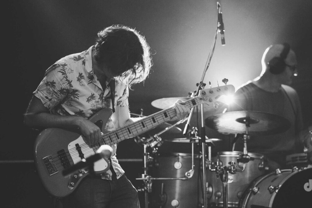 Ein Musiker spielt eine elektrische Bassgitarre auf der Bühne, mit gesenktem Kopf, neben einem Schlagzeuger mit Kopfhörern. Die schwarz-weiße Szene mit heller Bühnenbeleuchtung fängt die Energie ein - die perfekte Inspiration für jeden Webdesigner in Aachen.