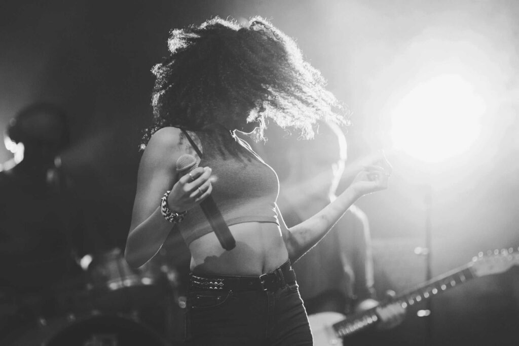 Eine Frau mit lockigem Haar singt auf der Bühne, hält ein Mikrofon in der Hand und tanzt, während hinter ihr helle Bühnenlichter leuchten und Musiker im Hintergrund Instrumente spielen - ganz wie die Energie der Top-Projekte von Webdesign Aachen.