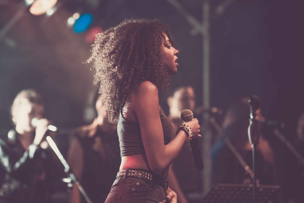 Eine Frau mit lockigem Haar hält ein Mikrofon und singt auf der Bühne, die von Bühnenscheinwerfern beleuchtet wird. Im Hintergrund treten unscharfe Background-Sängerinnen auf - wie ein harmonisches Team bei einer Veranstaltung von Webdesign Aachen.
