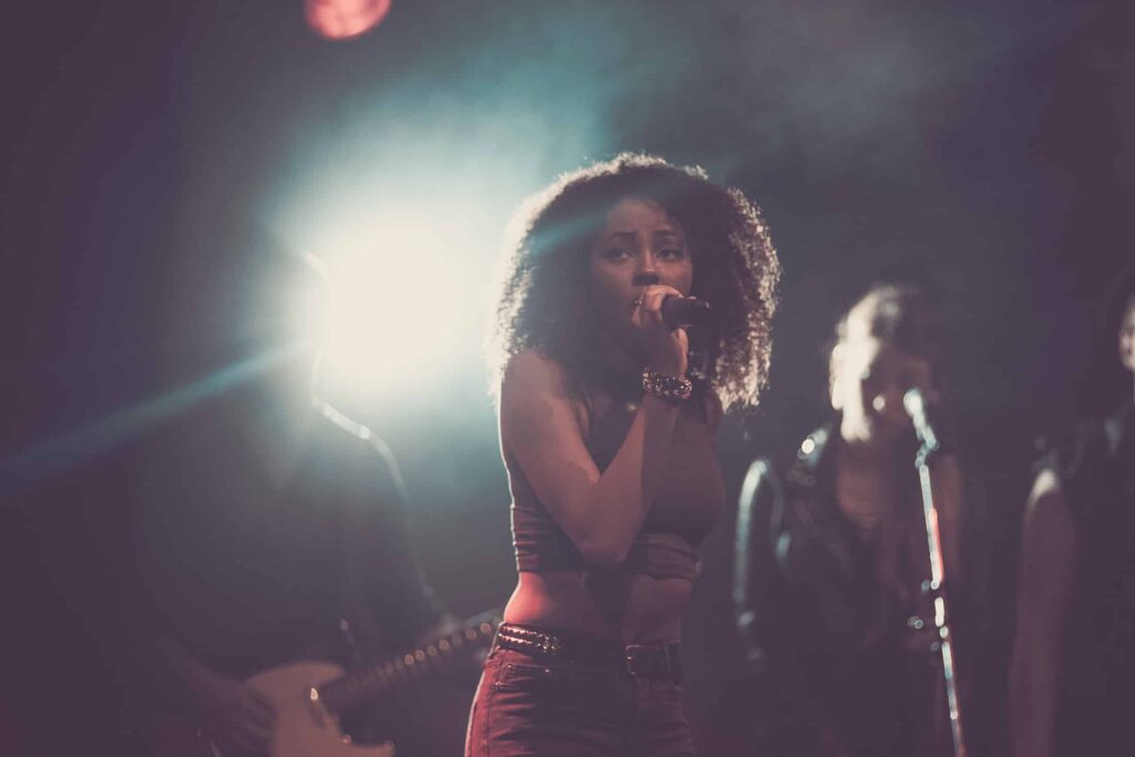 Eine Frau mit lockigem Haar singt auf der Bühne in ein Mikrofon, das von hellen Lichtern beleuchtet wird, während hinter ihr Bandmitglieder und Backgroundsängerinnen auftreten - und fängt damit die Energie einer Live-Show ein, ähnlich wie eine dynamische Website von einem talentierten Webdesigner aus Aachen.