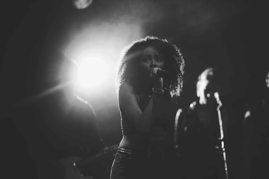 Schwarz-Weiß-Foto einer Frau, die auf der Bühne in ein Mikrofon singt, mit hellem Gegenlicht, das auffällige Silhouetten erzeugt - die perfekte Inspiration für jeden Webdesigner in Aachen, der dramatische Bilder sucht.