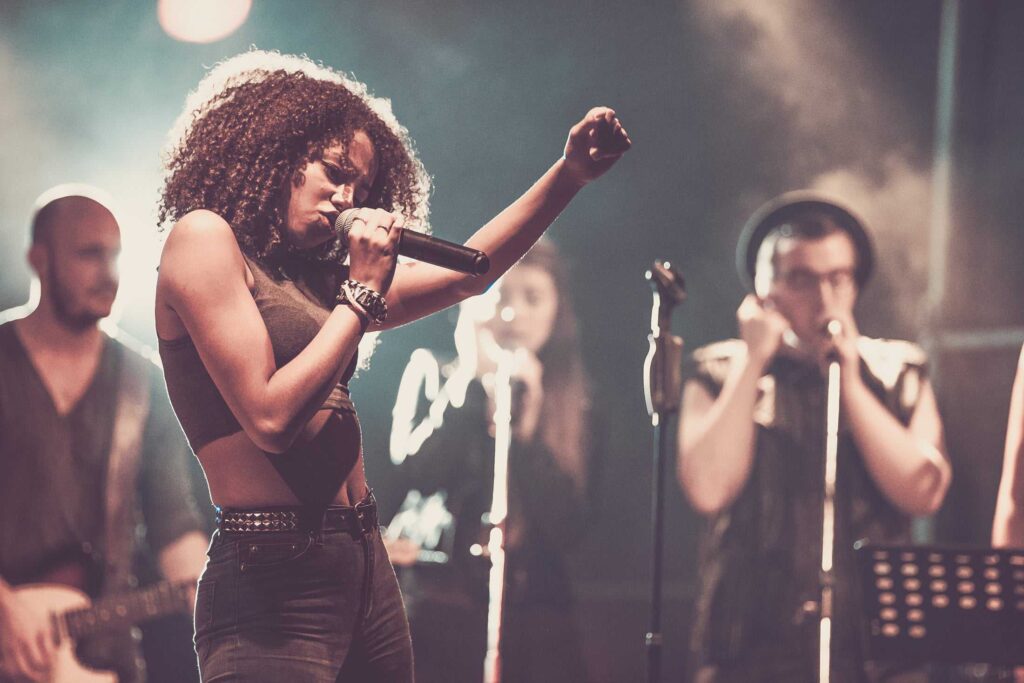 Eine Frau, die auf der Bühne mit erhobenem Arm leidenschaftlich in ein Mikrofon singt, während im Hintergrund eine Band und Backgroundsängerinnen bei dramatischer Beleuchtung auftreten - perfekt, um Ihre kreativen Projekte in einem Webdesign Aachen Portfolio zu präsentieren.