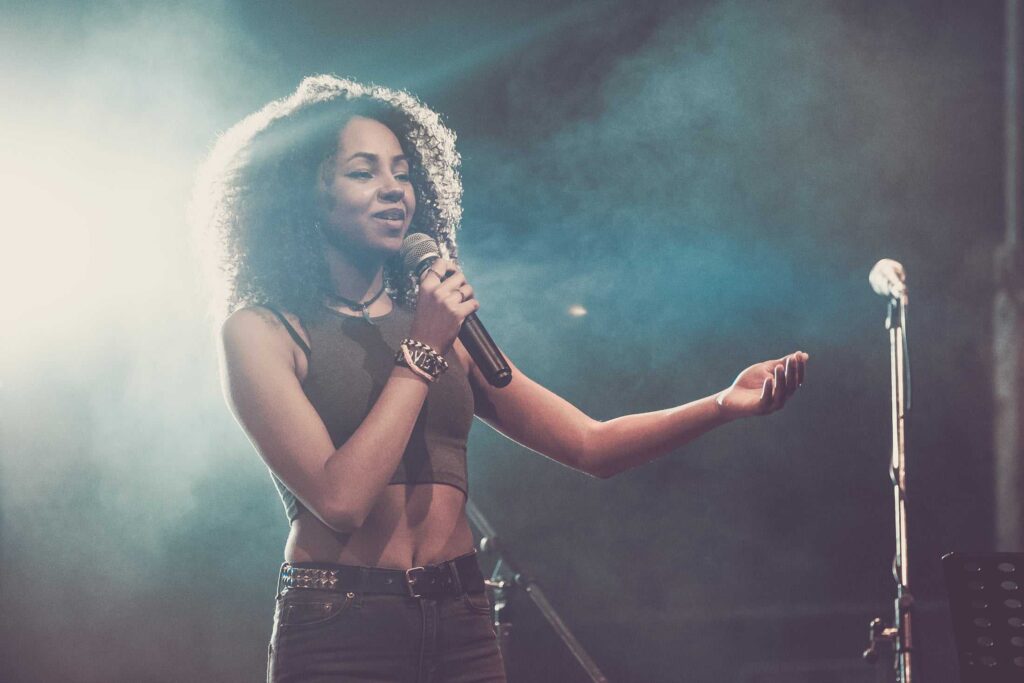 Eine Frau mit lockigem Haar singt auf einer verrauchten, schwach beleuchteten Bühne in ein Mikrofon. Sie trägt ein ärmelloses Crop-Top und Jeans, den Arm ausgestreckt - genau die Kreativität, die man von einem Top-Webdesigner aus Aachen erwartet.