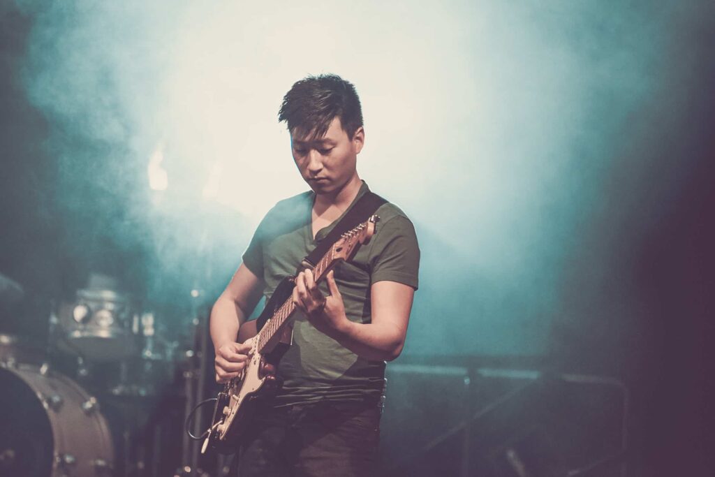 Ein Musiker spielt eine E-Gitarre auf der Bühne unter blauen und weißen Lichtern, mit einem Schlagzeug im Hintergrund und atmosphärischem Dunst - eine Kreativität, wie sie auch ein Webdesigner in Aachen in digitale Projekte einbringt.