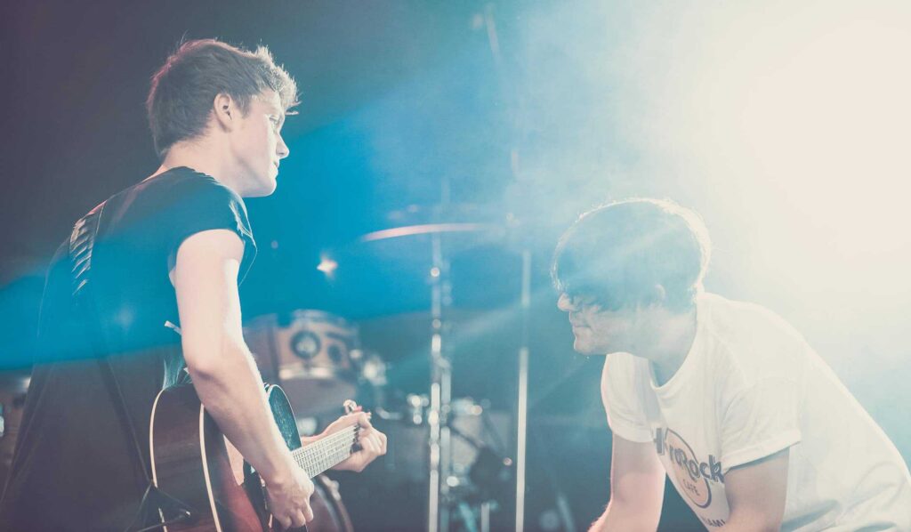 Zwei junge Männer treten auf der Bühne auf; einer spielt eine akustische Gitarre, während der andere kniet und singt. Beide werden von hellen Bühnenlichtern angestrahlt, während im Hintergrund die Musikanlage zu sehen ist. Das fängt den kreativen Geist ein - perfekt für jedes Webdesign Aachen Projekt.