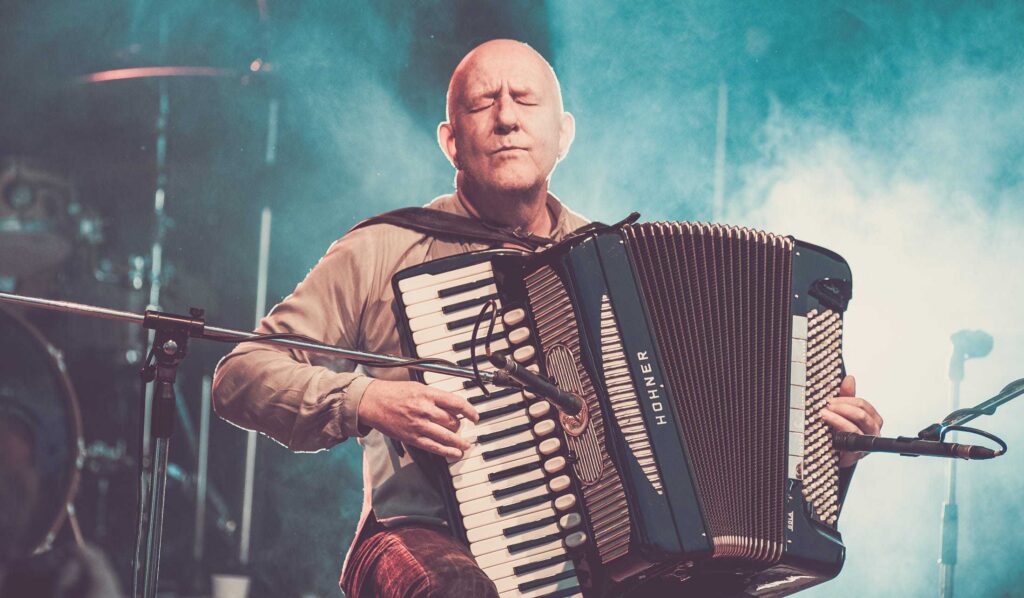 Ein glatzköpfiger Mann sitzt auf der Bühne und spielt mit geschlossenen Augen ein Akkordeon. Er scheint tief in die Musik versunken zu sein, während blaues Bühnenlicht und Nebel eine inspirierende Atmosphäre schaffen - ähnlich wie ein erfahrener Webdesigner in Aachen, der einzigartige digitale Erlebnisse schafft.