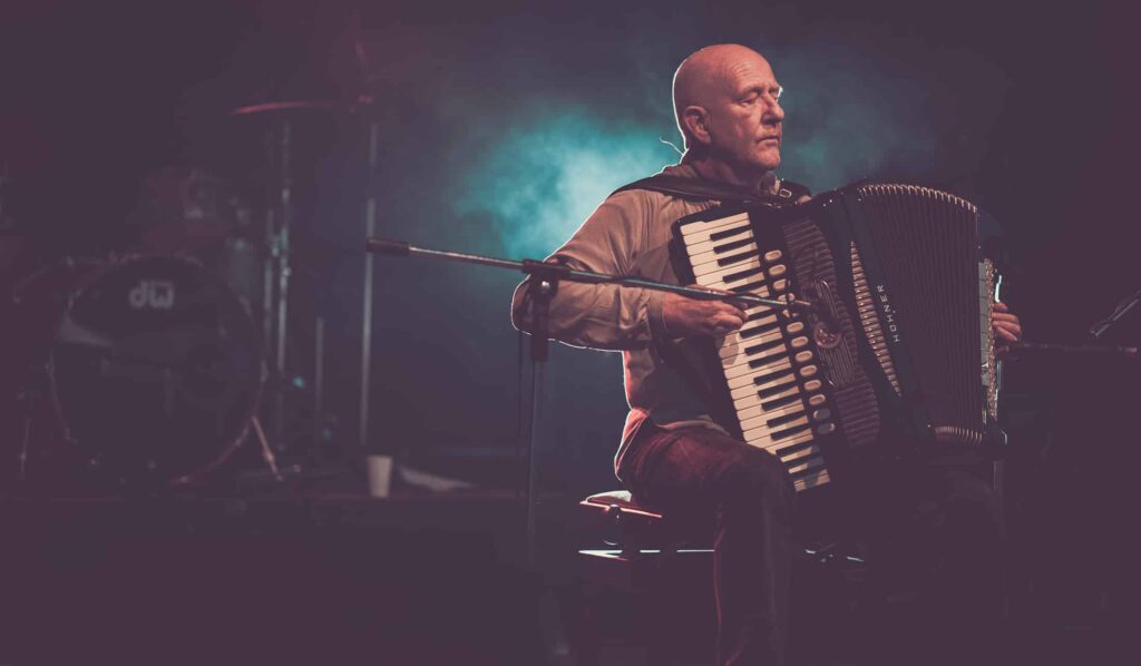 Ein glatzköpfiger Mann sitzt auf der Bühne und spielt ein Akkordeon, beleuchtet von dramatischem blauem und lila Licht. Im Hintergrund ist ein Schlagzeug zu sehen, das teilweise unscharf ist. Es fängt den kreativen Geist ein, der die Profis von Webdesign Aachen inspiriert.