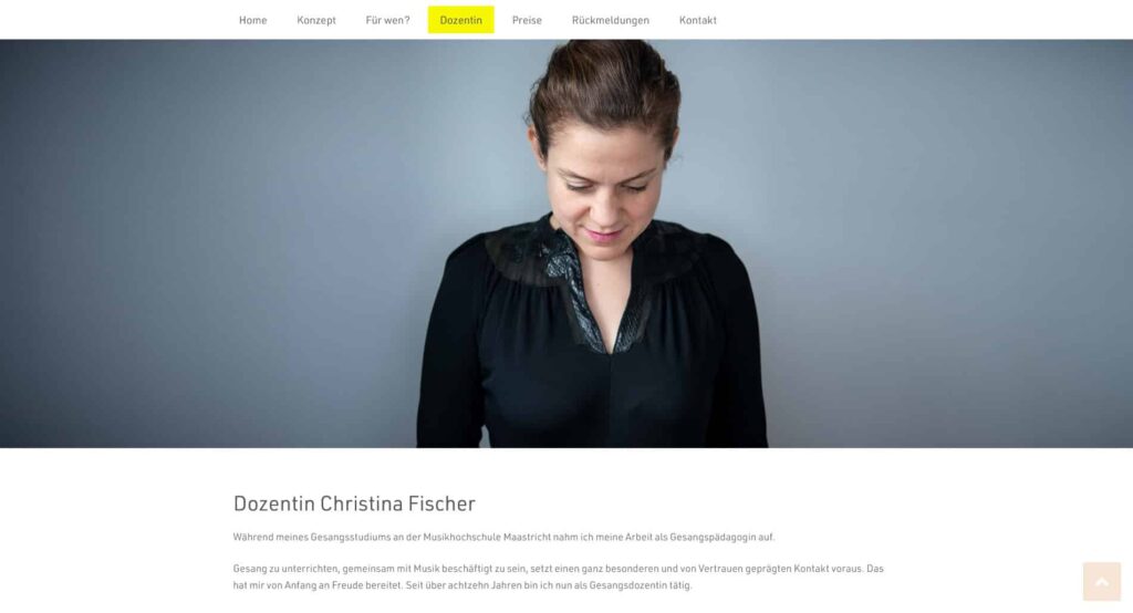 Eine Frau mit zurückgebundenen braunen Haaren, die eine schwarze Bluse mit Spitzendetails trägt, blickt mit einem leichten Lächeln auf einen schlichten grauen Hintergrund. Die Kopfzeile der Website und das Navigationsmenü von Webdesign Aachen sind über ihr zu sehen.