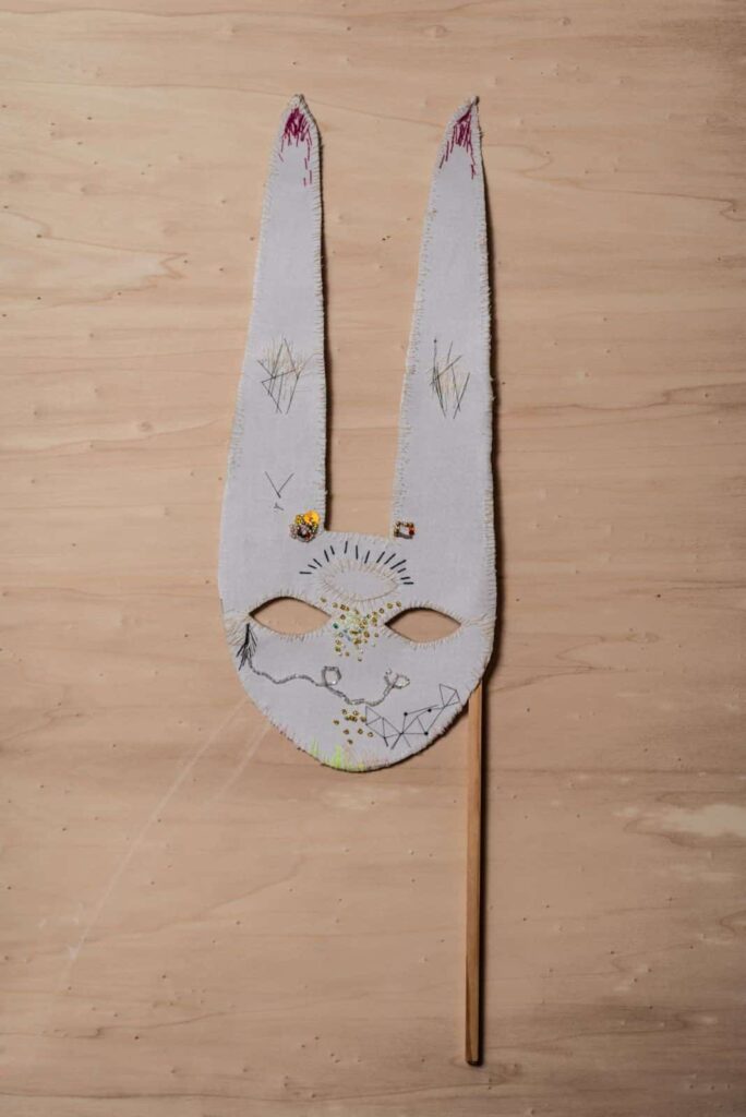 Eine handgefertigte Kaninchenmaske mit gestickten Details und zwei langen Ohren ist an einem Holzstab befestigt und liegt auf einer hellen Holzoberfläche - perfekt für kreative Inspiration, egal ob Sie sich für Basteln oder Webdesign im Aachener Stil interessieren. Die Maske ist mit einfachen Stickereien und dekorativen Elementen versehen.