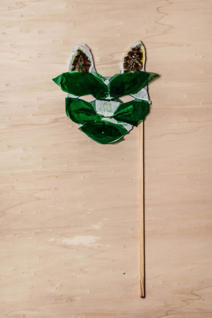 Eine handgefertigte Tiermaske aus grünem und weißem Papier, geformt wie ein Gesicht mit zwei Ohren und befestigt an einem Holzstab, liegt auf einer hellen Holzfläche - die perfekte Inspiration für kreative Webdesign-Projekte in Aachen.
