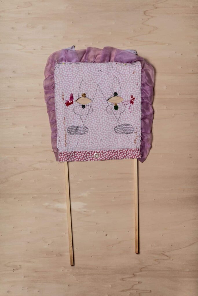 Ein handgefertigtes Stoffschild mit einer rosa Rüschenbordüre, auf dem zwei abstrakte gestickte Figuren - jede mit einem Kreis als Kopf - auf Holzstäben montiert sind, wird vor einem hellen Holzhintergrund ausgestellt, perfekt für jedes kreative Webdesigner-Aachen-Studio.