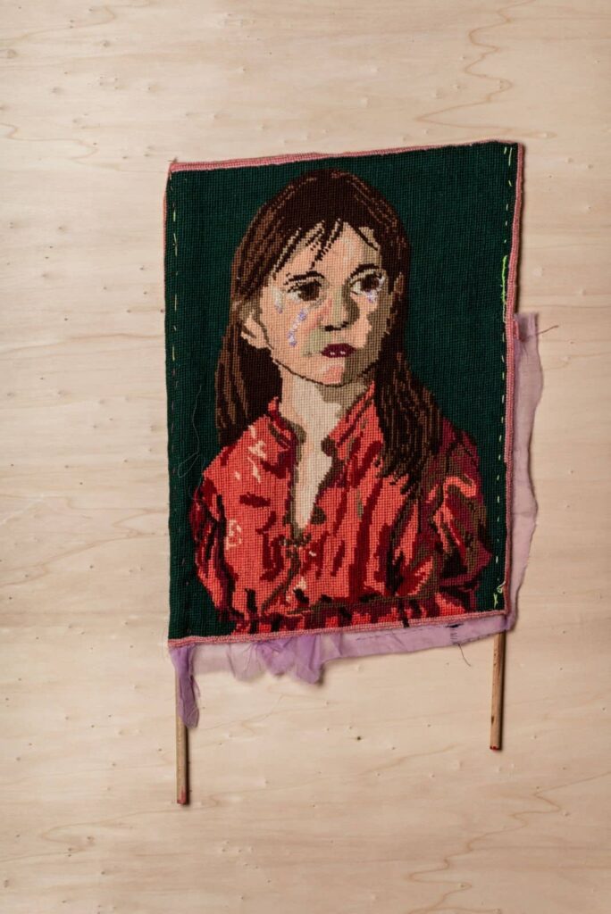 Ein textiles Kunstwerk auf Stäben zeigt ein junges Mädchen mit braunen Haaren und einem roten Hemd vor einem dunkelgrünen Hintergrund. Die perfekte Inspiration für jedes Webdesign Aachen Projekt. Der Stoff hat ausgefranste Ränder und ist auf einer hellen Holzoberfläche ausgestellt.