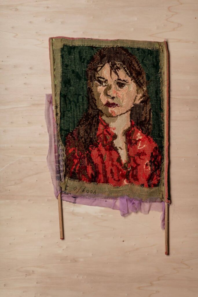 Ein gewebtes Textilporträt eines Mädchens mit langen braunen Haaren und einem rot gemusterten Hemd vor grünem Hintergrund wird auf zwei Holzstäben mit durchscheinendem lila Stoff an den Rändern präsentiert - die perfekte Inspiration für jedes Webdesign Aachen-Projekt.