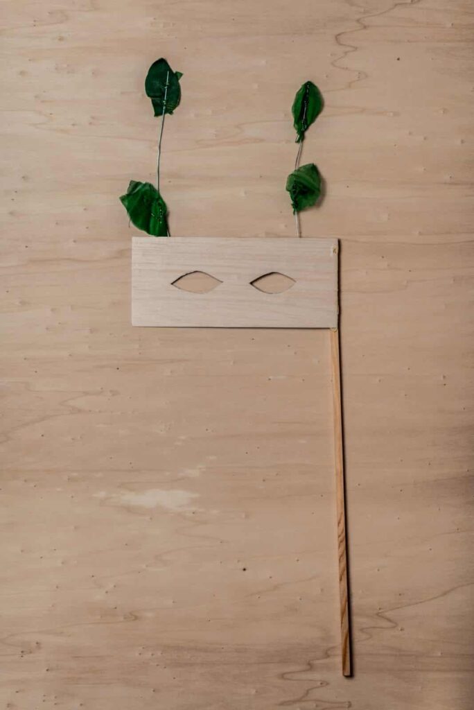 Eine einfache Maske aus einem rechteckigen Holzstück mit zwei Augenlöchern, die an einem Holzstab befestigt und mit zwei grünen Blättern an Drahtstielen verziert ist, die an Antennen erinnern. Ideal als Inspiration für kreative Webdesign Aachen Projekte.