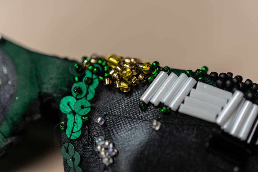 Nahaufnahme einer dekorativen Perlenarbeit auf Stoff mit grünen Pailletten, gelben und goldenen runden Perlen, weißen zylindrischen Perlen und schwarzen Akzentperlen, die in einem Muster angeordnet sind - eine ideale Inspiration für kreative Webdesign Aachen Projekte.