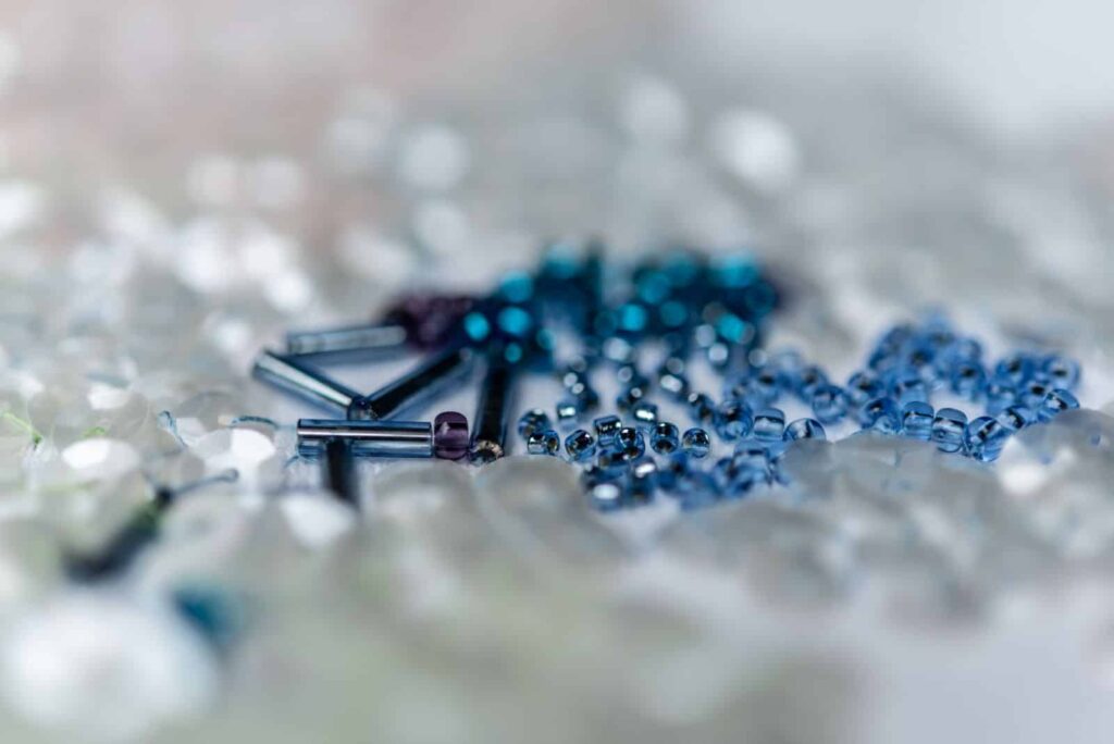 Eine Nahaufnahme verschiedener kleiner Perlen, darunter durchsichtige, blaue und zylindrische Metallperlen, die auf einer hellen Oberfläche mit einem weichen, unscharfen Hintergrund verstreut sind - perfekt für kreative Projekte oder inspirierende Ideen für Webdesign Aachen.
