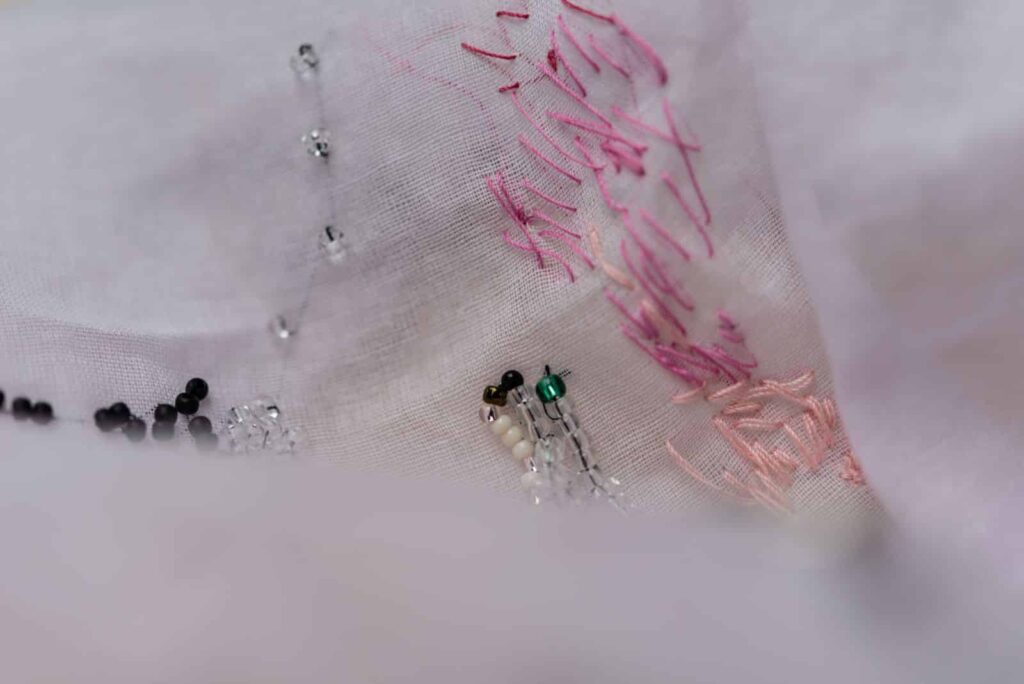 Nahaufnahme eines hellen Stoffes mit rosa, weißen und schwarzen Stickereistichen und kleinen Perlen, darunter eine grüne Perle, die ein abstraktes, strukturiertes Design ergeben - die perfekte Inspiration für jeden kreativen Webdesigner Aachen.