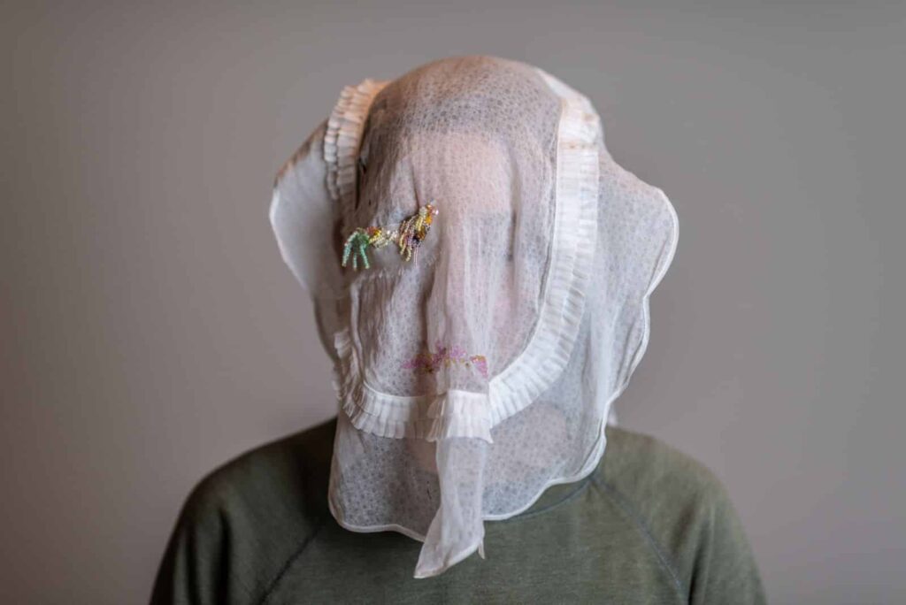 Eine Person, die ein grünes Hemd trägt und deren Kopf von einem weißen, strukturierten, mit Perlen verzierten Tuch bedeckt ist, das ihr Gesicht verdeckt, steht vor einem schlichten grauen Hintergrund - ein faszinierendes Bild für jedes Webdesigner Aachen Projekt.