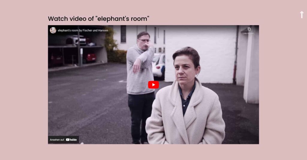 Eine Frau in einem hellen Mantel steht im Vordergrund und schaut ernst, während ein Mann in einem grauen Pullover hinter ihr auf einem Parkplatz gestikuliert. Das angehaltene YouTube-Video deutet auf eine Projektbesprechung von Webdesign Aachen hin.