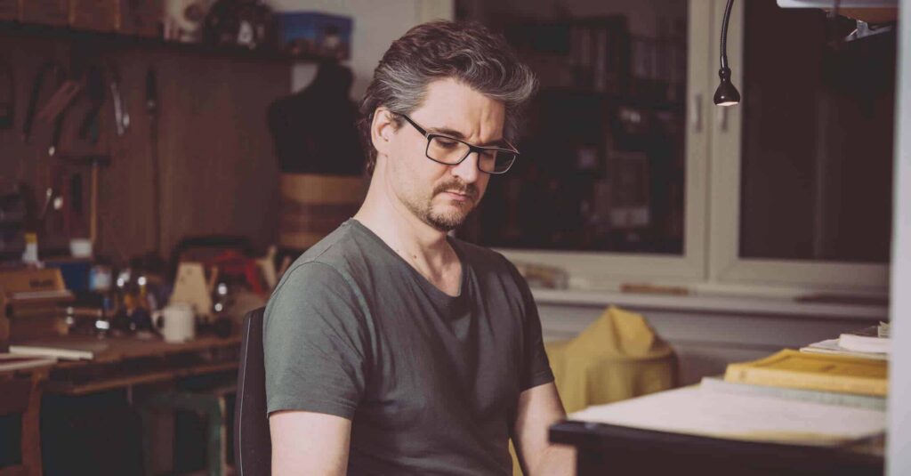 Ein Mann mit Brille und kurzen dunklen Haaren, der ein graues T-Shirt trägt, sitzt drinnen an einem Schreibtisch und blickt nachdenklich nach unten - vielleicht plant er sein nächstes Webdesign Aachen-Projekt -, während im Hintergrund Regale und Werkzeuge zu sehen sind.