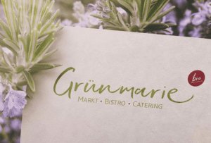 Ein Schild mit der Aufschrift "Gr&uuml;nmarie MARKT - BISTRO - CATERING" in gr&uuml;ner und roter Schrift und einem kleinen roten Kreis mit der Aufschrift "bio" liegt inmitten von Lavendel- oder Rosmarinzweigen - die perfekte Inspiration f&uuml;r ein Webdesign-Projekt in Aachen.