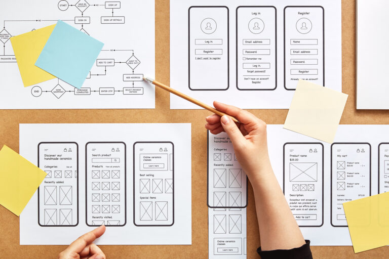 Man sieht Hände, die an auf einem Tisch ausgebreiteten Prototypen für mobile App-Designs arbeiten. Die Blätter zeigen Anmeldebildschirme, Suchoberflächen und Inhaltslayouts. Haftnotizen und Flussdiagramme sind ebenfalls vorhanden. Ein Aachener Webdesigner zeigt mit einem Bleistift auf ein Drahtmodell.