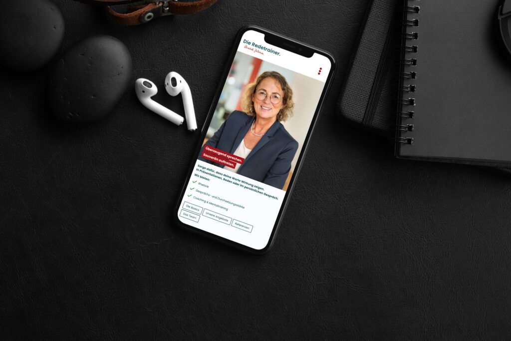 Ein Smartphone auf einer schwarzen Lederoberfläche zeigt das Profil einer professionellen Frau. In der Nähe befinden sich kabellose Ohrhörer, glatte schwarze Steine, ein braunes Uhrenarmband und ein Spiralnotizbuch mit einem Stift.