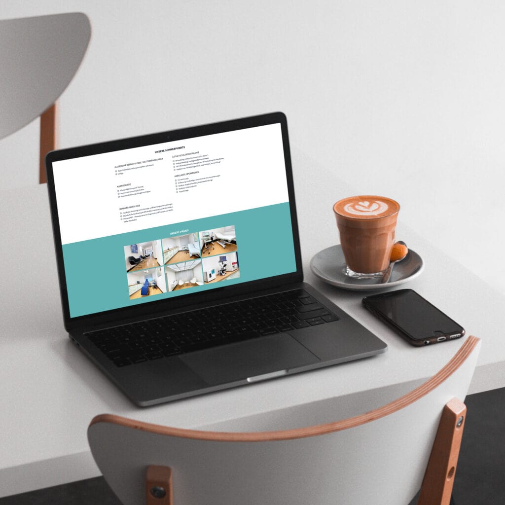 Ein Laptop mit einer Website steht auf einem weißen Tisch neben einem Cappuccino mit Latte Art, einer Untertasse und einem Smartphone; daneben steht ein weißer Stuhl.