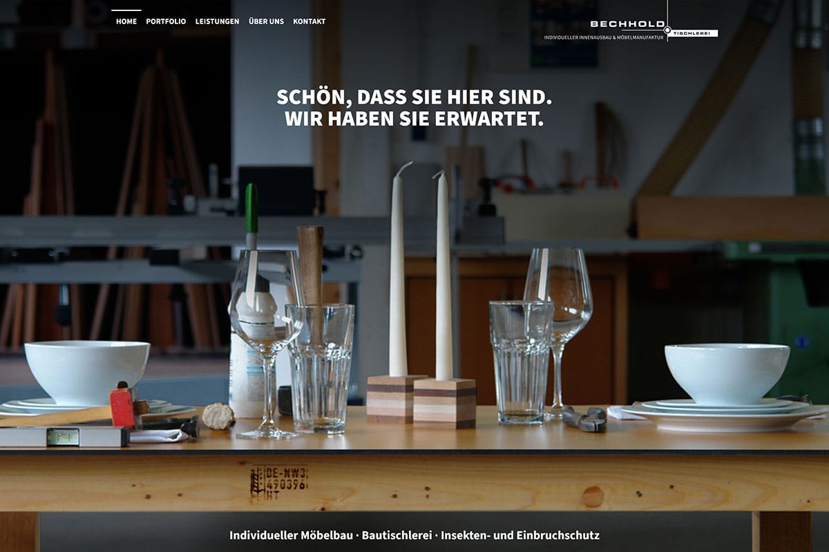 Ein gedeckter Esstisch mit weißen Tellern, Gläsern und Silberbesteck steht im Vordergrund einer Holzwerkstatt, in der im Hintergrund Holzregale, Werkzeuge und Geräte zu sehen sind. Über dem Tisch erscheint ein deutscher Text.